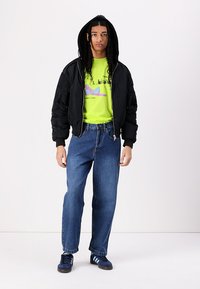 Black bomber jacket with hood, neon green graphic t-shirt, loose blue jeans, and navy sneakers with white stripes.
