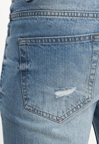 Jeans de mezclilla azul claro con un bolsillo trasero que presenta un detalle desgastado. La tela tiene una textura suave y costuras visibles.