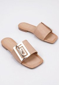 Sandalias deslizantes beige con punta cuadrada, tacón plano y una gran hebilla geométrica metálica en la correa superior. Textura suave y diseño minimalista.
