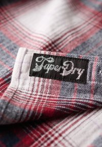 Tissu à carreaux rouge, gris et blanc avec une texture douce. Présente une étiquette noire avec le texte argenté "SuperDry" au niveau du col.