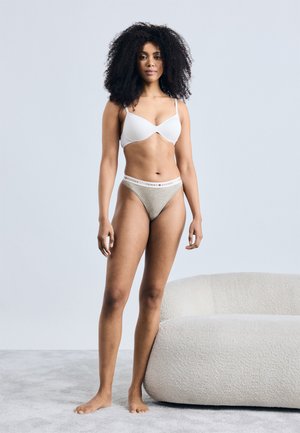 Reggiseno wireless bianco in tessuto liscio con spalline sottili, abbinato a perizoma beige con fascia logo. Sfondo neutro e seduta imbottita.