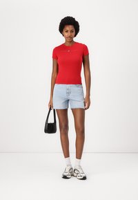 Rød kortermet tettsittende t-skjorte med tekstdetalj, kombinert med lyse denimshorts, hvite sokker og svart håndveske, fullført med sølvfargede joggesko.