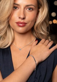 Silver bracelet on wrist, close-up of a disc pendant necklace, smooth skin, blonde hair, dark blue fabric top, soft focus background lights.