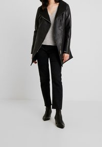 Schwarze Lederjacke mit breitem Kragen, Reißverschluss an der Seite und asymmetrischem Saum. Getragen über einem grauen V-Ausschnitt-Oberteil und schwarzen Jeans.