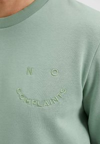 Detailní záběr na světle zelenou mikinu s výšivkou textu "NO COMPLAINTS" tvořícího obličej s úsměvem na hrudi.