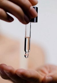 Bottiglia di vetro trasparente con tappo nero, contenente un liquido chiaro; contagocce che rilascia una goccia sopra un palmo. Superficie liscia.