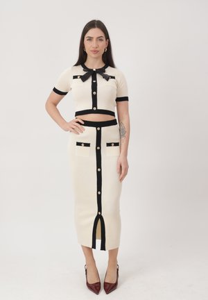 Frau trägt ein cremefarbenes zweiteiliges Outfit mit schwarzem Besatz, goldenen Knöpfen, einem Oberteil mit Schleife, einem langen Rock mit Taschen und burgunderroten Absätzen, stehend.