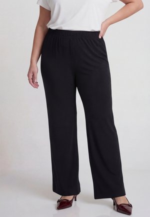 Kvinde iført sorte bukser med vide ben, hvid kortærmet skjorte som er stukket ind, og burgunderrøde spidse hæle med lav hæl, stående med hånden på hoften.