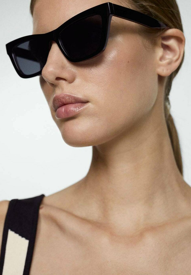 Zwarte cat-eye zonnebril met een dikke montuur en donkere lenzen, die een gedurfde, hoekige uitstraling toont. Glanzende textuur aanvult de stijlvolle look.