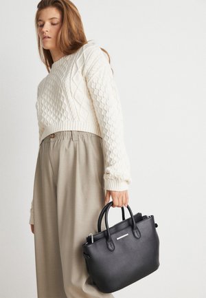 Frau trägt einen weißen Zopfpullover und beige weite Hosen, hält eine schwarze Ledertasche mit zwei Griffen.