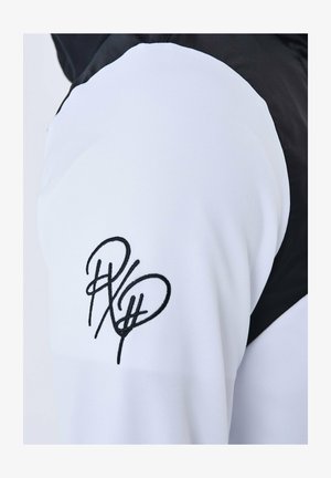 Veste blanche et noire avec un tissu texturé, présentant un logo noir brodé en relief sur la manche et un design en blocs de couleur.