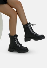 Botas de combate de cuero negro con suela gruesa, que presentan detalles en plata en los cordones y una cremallera en la parte delantera. Acabado texturizado con diseño de cordones.
