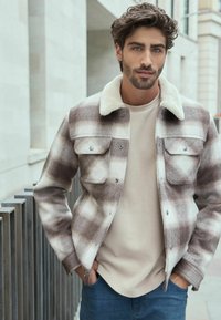 Brown and cream plaid jacket with a shearling collar, two chest pockets, and a light beige shirt underneath. Paired with blue denim jeans.