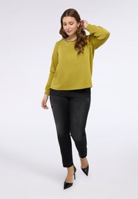 Olivově zelená mikina se zdobnými kamínky na límci, spárovaná s roztrhanými černými džíny a černými vysokými podpatky. Hladká textury látky.