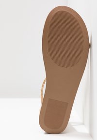 Semelle en caoutchouc marron d'une sandale, présentant un motif texturé. La partie supérieure est en matériau beige clair avec de fines sangles.