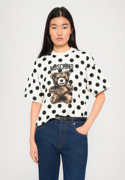 Person wearing white oversized polka dot t-shirt with teddy bear and "MOSCHINO" text, paired with blue jeans and Moschino belt.