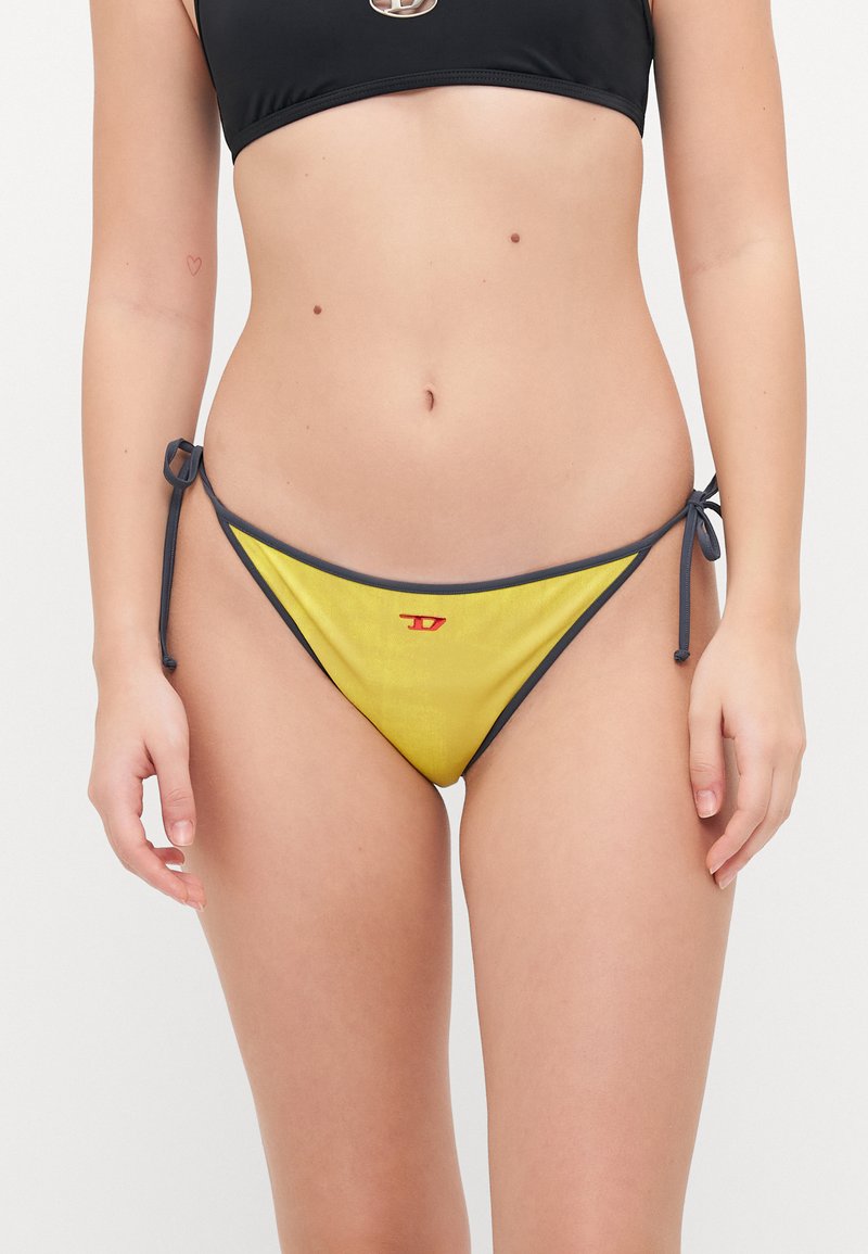 Gul bikini-bunn med mørk grå kant, sidesnøring og en liten rød logo på forsiden. Glatt tekstur og trekantet form.