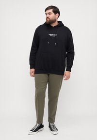Svart hettegenser med en frontlomme og tekstdetalj, kombinert med olivengrønne bukser og svarte joggesko med hvite detaljer.