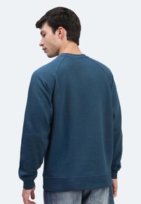 Uomo con capelli scuri che indossa un semplice maglione blu a maniche lunghe e jeans blu sbiaditi, girato leggermente di lato su uno sfondo semplice.