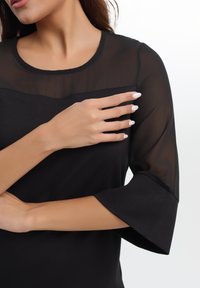 Črna majica z prosojnimi rokavi, okroglo ovratnikom in mehkobo. Rokavi so razširjeni, material pa ima gladek zaključek.
