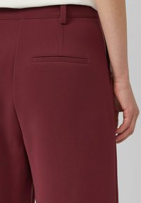 Pantalons bordeaux avec une texture lisse, présentant un design classique et une poche arrière unique. La taille est dotée d'une passante de ceinture.