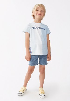 Camiseta de algodón de color azul claro con la impresión "SALVA LOS OCÉANOS", combinada con pantalones cortos de denim. El calzado son zapatillas amarillas tipo slip-on con suela blanca.