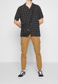 Schwarzes Kurzarmhemd mit weißem geometrischem Muster, khakifarbene Cargo-Hose mit Seitentaschen und schwarze Sneakers mit weißen Sohlen.