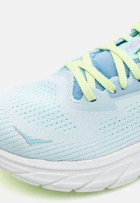 Světle modrá běžecká bota s síťovaným svrškem, texturovaným designem a neonově zelenými tkaničkami. Má polstrovanou bílou podrážku a růžový akcentovaný logo.