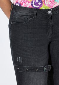 Jean en denim noir avec une texture délavée, présentant une zone déchirée et une sangle avec des accessoires en métal à l'ourlet.