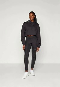 Čierna skrátená mikina s kapucňou a šnúrkami, kombinovaná s čiernymi legínami s vysokým pásom a jemnými akcentmi. Model má na sebe biele tenisky.