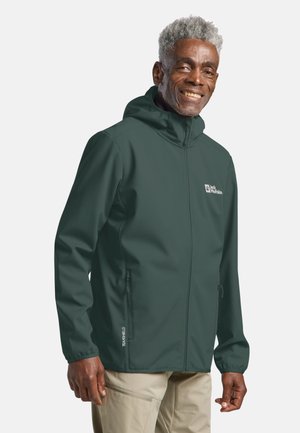 Älterer Mann, der eine dunkelgrüne Kapuzenjacke mit Reißverschluss und beige Hosen trägt, lächelnd und vor einem einfarbigen Hintergrund steht.