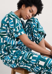 Tvådelad outfit i teal, vitt och grönt geometriskt mönster. Avslappnad passform, korta ärmar, lösa byxor med en mjuk textur. Bakgrund av trästol.