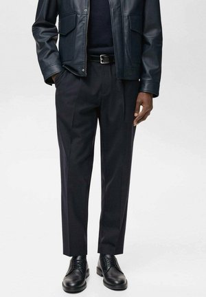 Homme portant une veste en cuir noire, une ceinture noire, un pantalon noir sur mesure et des chaussures habillées noires et brillantes, debout devant un fond uni.