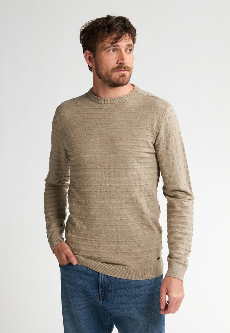 Beige gestrickter Pullover mit strukturierten horizontalen Streifen, Rundhalsausschnitt und gerippten Bündchen, kombiniert mit blauen Jeans.