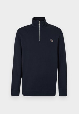 Mladý muž stojaci s jednou rukou v vrecku, oblečený v námorníckom modrom pulóvri so zipsom do polovice, sivých plisovaných nohaviciach a bielych teniskách.