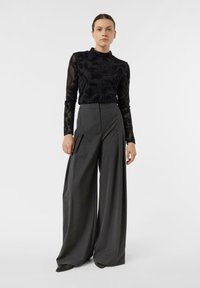 Kobieta stojąca, ubrana w czarną przezroczystą bluzkę z długim rękawem i wzorem paisley oraz szare, szerokie spodnie z wysokim stanem na białym tle.