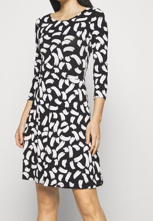 Kobieta w czarnej sukience z białym abstrakcyjnym wzorem pędzla i rękawami do połowy łokcia, stojąca na jednolitym tle.