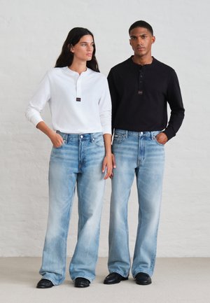 Deux modèles posent côte à côte, vêtus de hauts blancs et noirs à manches longues, de jeans bleu clair à jambes larges et de chaussures noires, devant un mur de studio uni.