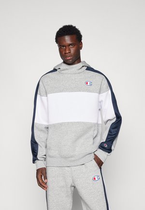 Sudadera gris y blanca con capucha, que presenta un logo de Champion, acentos laterales en navy y un corte relajado. Textura suave y puños de canalé.