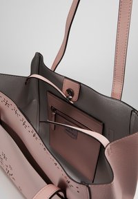 Różowa skórzana torba typu tote o gładkiej fakturze, z wewnętrzną kieszenią na zamek oraz dekoracyjnymi perforacjami. Posiada długie, płaskie rączki oraz zapięcie na zatrzask.