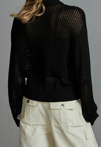 Schwarzer Strickpullover mit einem Mesh-Design und geripptem Saum, kombiniert mit elfenbeinfarbenen Cargo-Hosen mit Klappentaschen und Druckknöpfen.