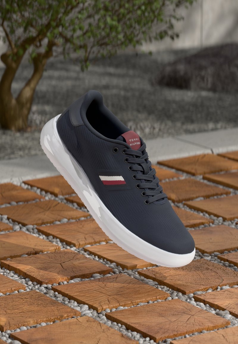 Basket bleu marine avec détails de rayures blanches et rouges, semelle blanche, posée sur des dalles en bois avec des espaces remplis de gravier, environnement extérieur avec un arbre en arrière-plan.