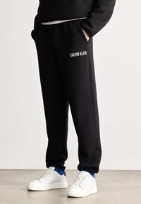 Czarne dresy z elastycznym pasem, bocznymi kieszeniami oraz białym logo "CALVIN KLEIN" na lewym udzie, w połączeniu z białymi sneakersami.