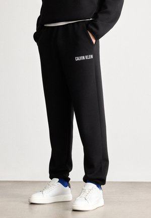 Musta verkkarit joustavyötäröllä, sivutaskuilla ja valkoisella "CALVIN KLEIN" -logolla vasemmassa reidessä, yhdistetty valkoisiin tennareihin.