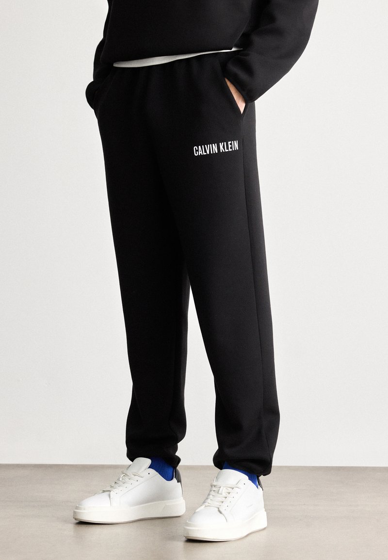 Czarne dresy z elastycznym pasem, bocznymi kieszeniami oraz białym logo "CALVIN KLEIN" na lewym udzie, w połączeniu z białymi sneakersami.