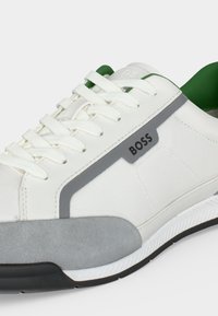 Baskets blanches avec lacets blancs, détail en daim gris, semelle noire, doublure intérieure verte et marquage "BOSS" sur la tranche.