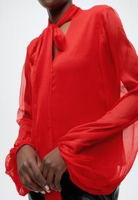 Blouse en chiffon rouge avec un col à nouer, manches longues et poignets froncés. Texture transparente avec une coupe fluide et une finition lisse.