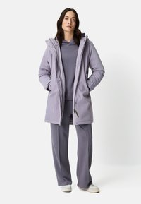 Lichtpaarse waterdichte jas met capuchon, gecombineerd met een bijpassende top met lange mouwen en wijde broekspijpen, gedragen met witte sneakers.