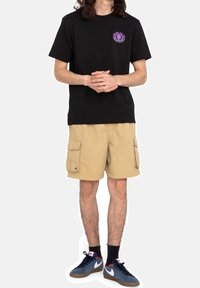 T-shirt noire avec logo violet, shorts cargo kaki avec poches, et baskets bleues avec accents blancs, se tenant devant un fond uni.