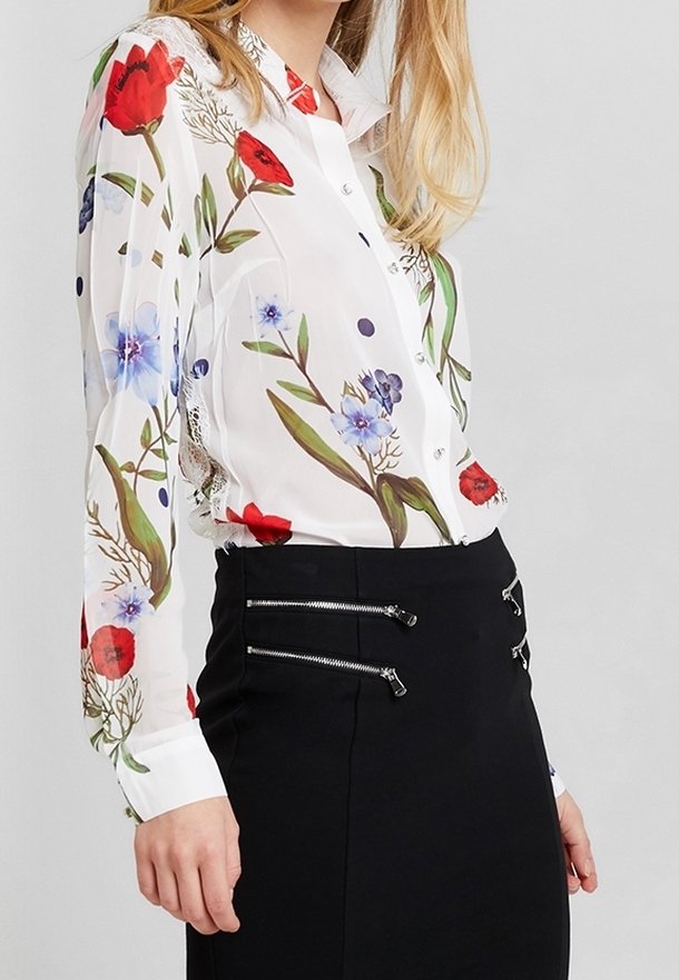 Blouse blanche à manches longues avec imprimé floral rouge et bleu, associée à une jupe noire avec plusieurs fermetures éclair horizontales argentées.